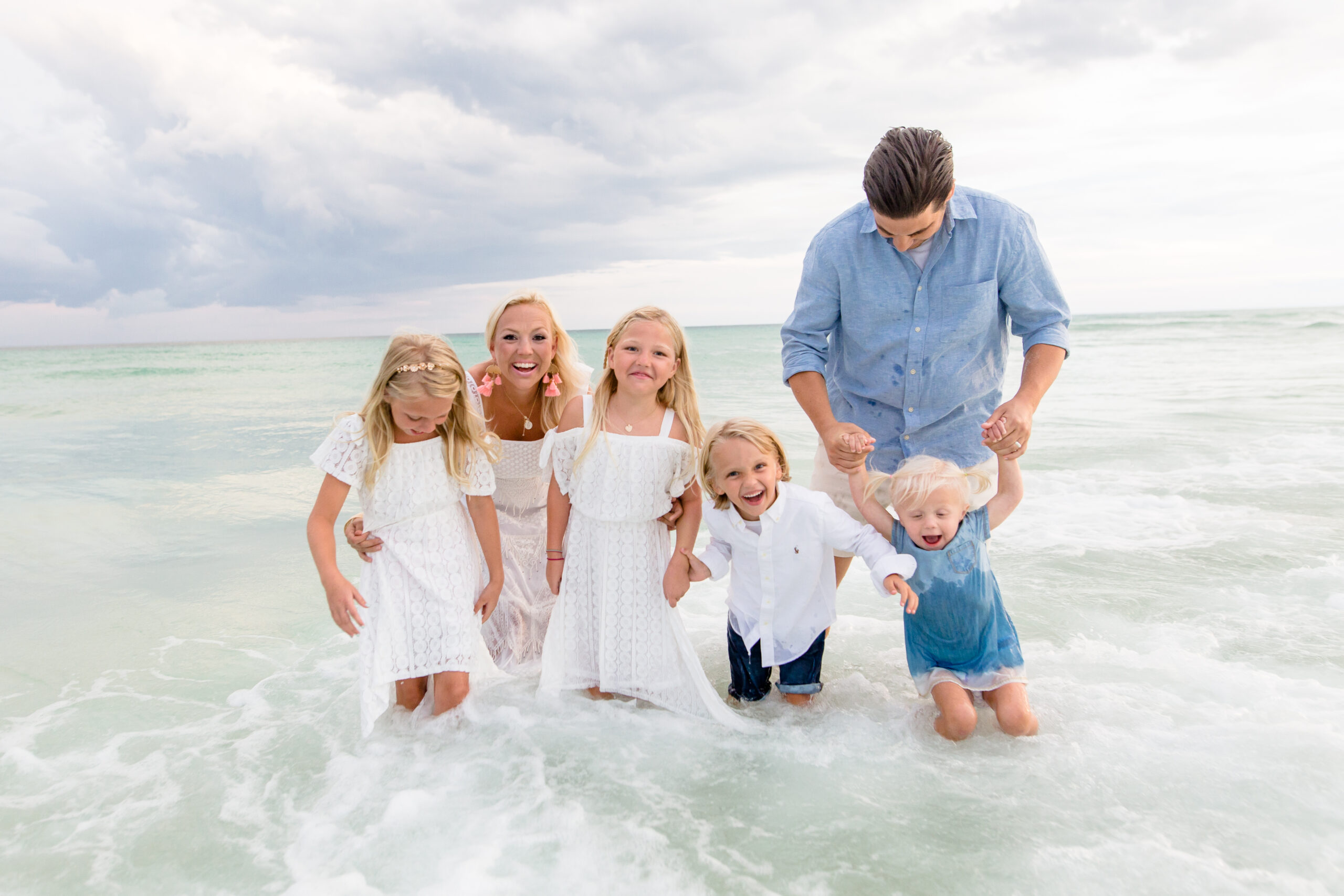 A family of six stands and plays in the shallow water at a beach, with waves splashing around them. The group includes two adults and four children, all smiling and wearing white and blue outfits.