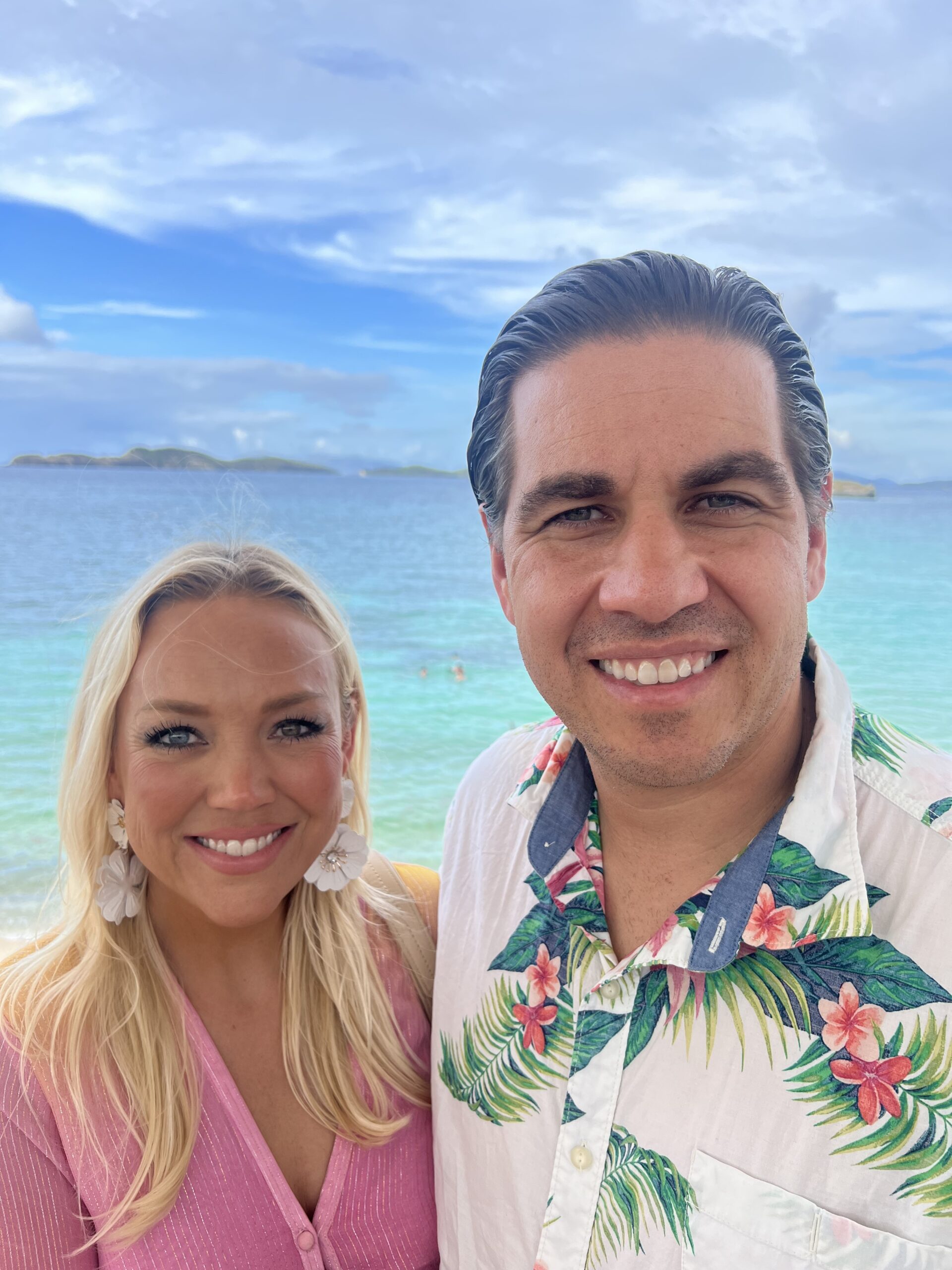 A woman in a pink top and a man in a white shirt with a tropical design stand by the ocean on a sunny day.