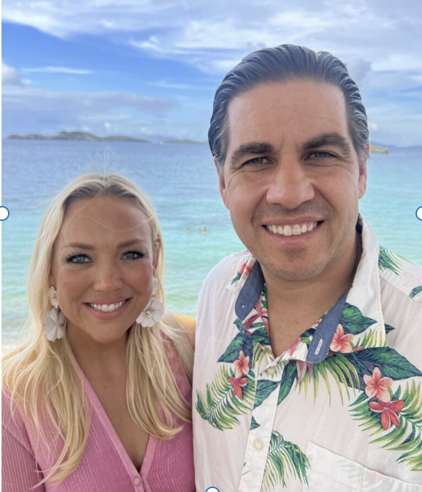 A woman in a pink top and a man in a white shirt with a tropical design stand by the ocean on a sunny day.